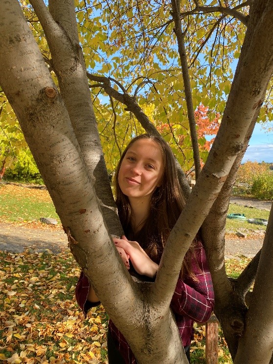 Woman wearing a maroon flannel shirt smiling while posing in between two tree branches on a fall day.
