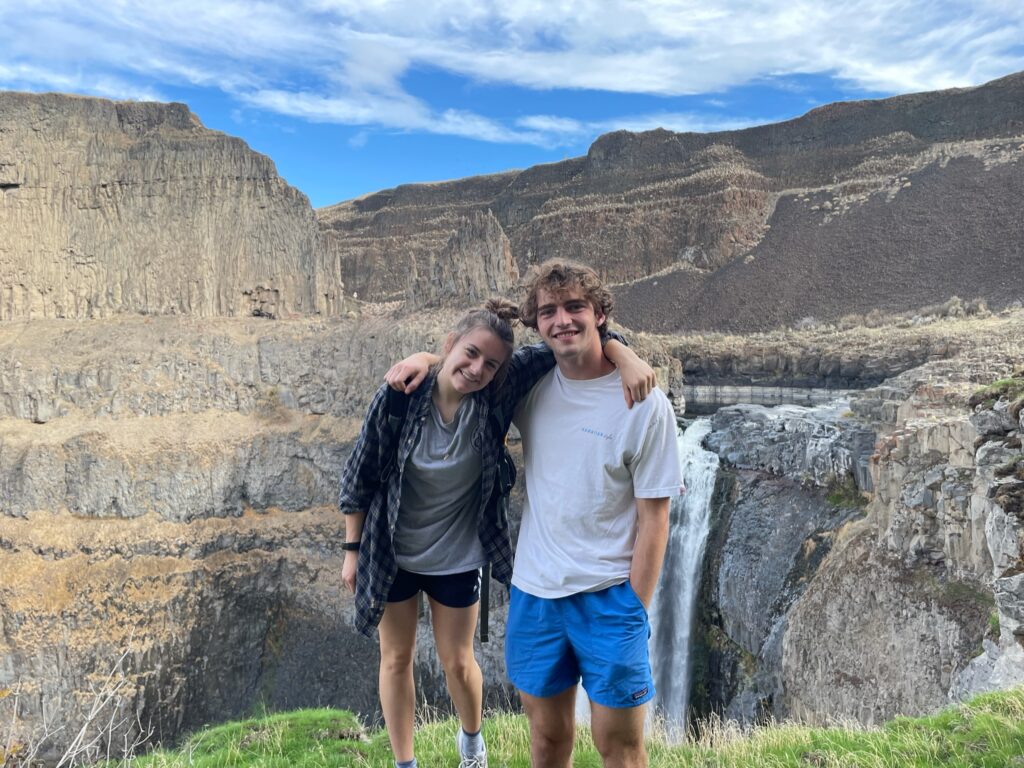 man and woman standing side by side with their arms around each other standing in front of  large mountains with a waterfall. 