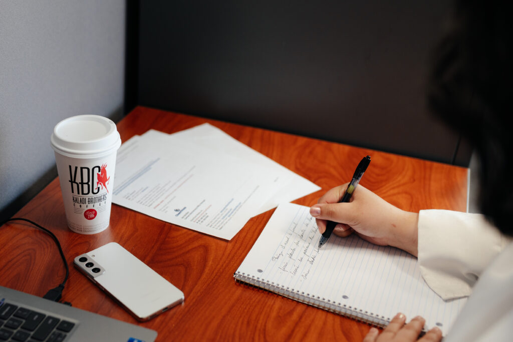 hand of a woman writing on a note pad. On the same table is a Kaldi Brothers coffee cup and documents.