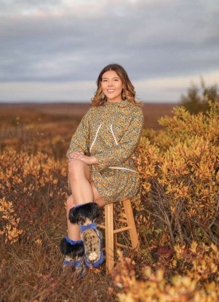 Woman sitting on a stool wearing  kuspuk and mukluks smiling, surrounded by yellow bushes in Kotzebue, Alaska.