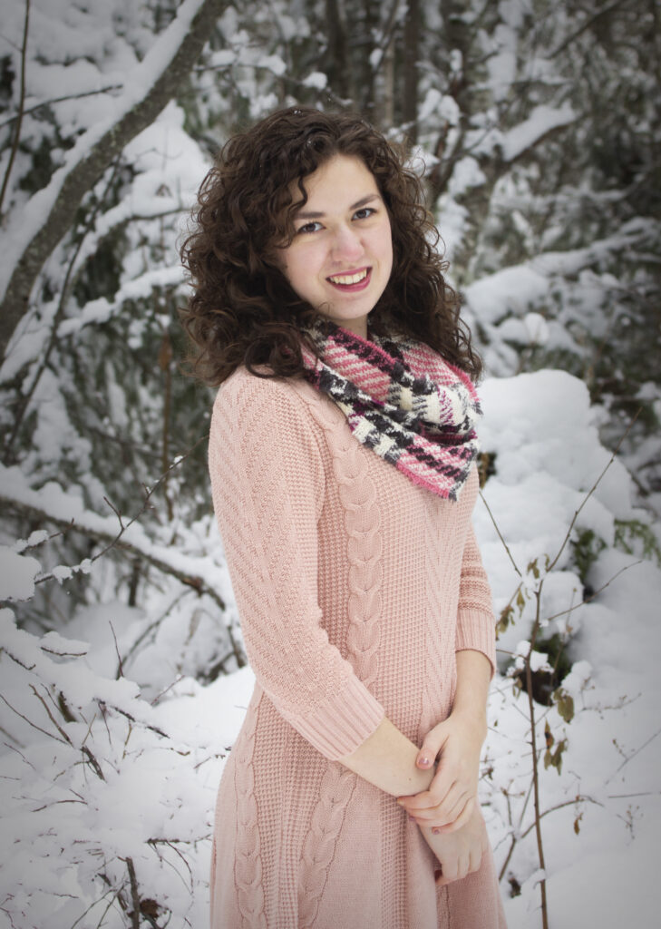 Woman in pink dress and black and pink scarf smiling in front of snow covered trees. 