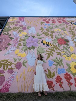 Woman with a long yellow dress standing in front of a pink wall painted with flowers and bees. 