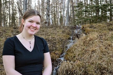 Woman in black t shirt smiling in front of a small stream on a fall day. 