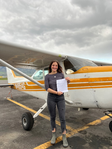 A Woman standing in front of a yellow propeller plane holding a certificate and smiling. 