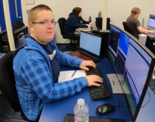Man wearing a blue sweatshirt and glasses sitting in front of a computer monitor smirking. 